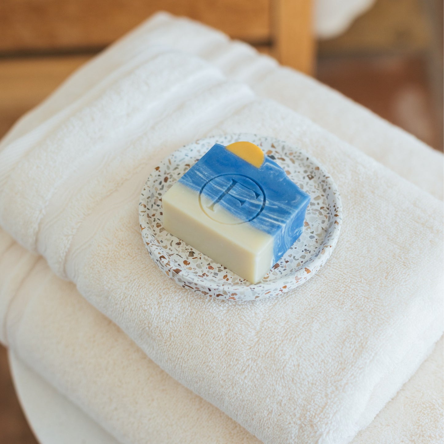 Savon artisanal FLAMARDA posé sur une coupelle en terrazzo, avec un motif océan bleu et soleil couchant, délicatement posé sur un drap de bain sable naturel. Savon fait main, esthétique et sensoriel, idéal pour une salle de bain élégante.