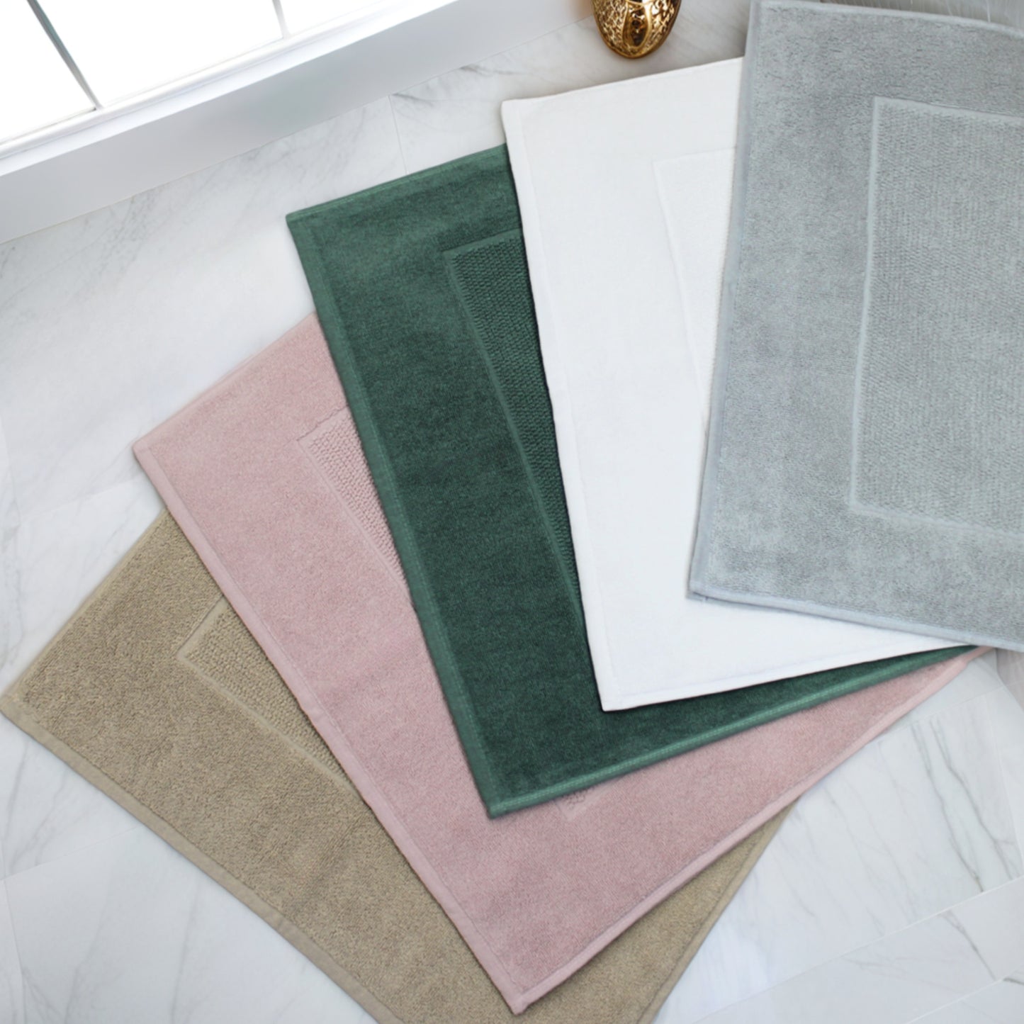 Tapis de bain FLAMARDA haut de gamme en différentes teintes douces, posés sur sol en marbre près d'une fenêtre, illustrant confort et élégance dans la salle de bain