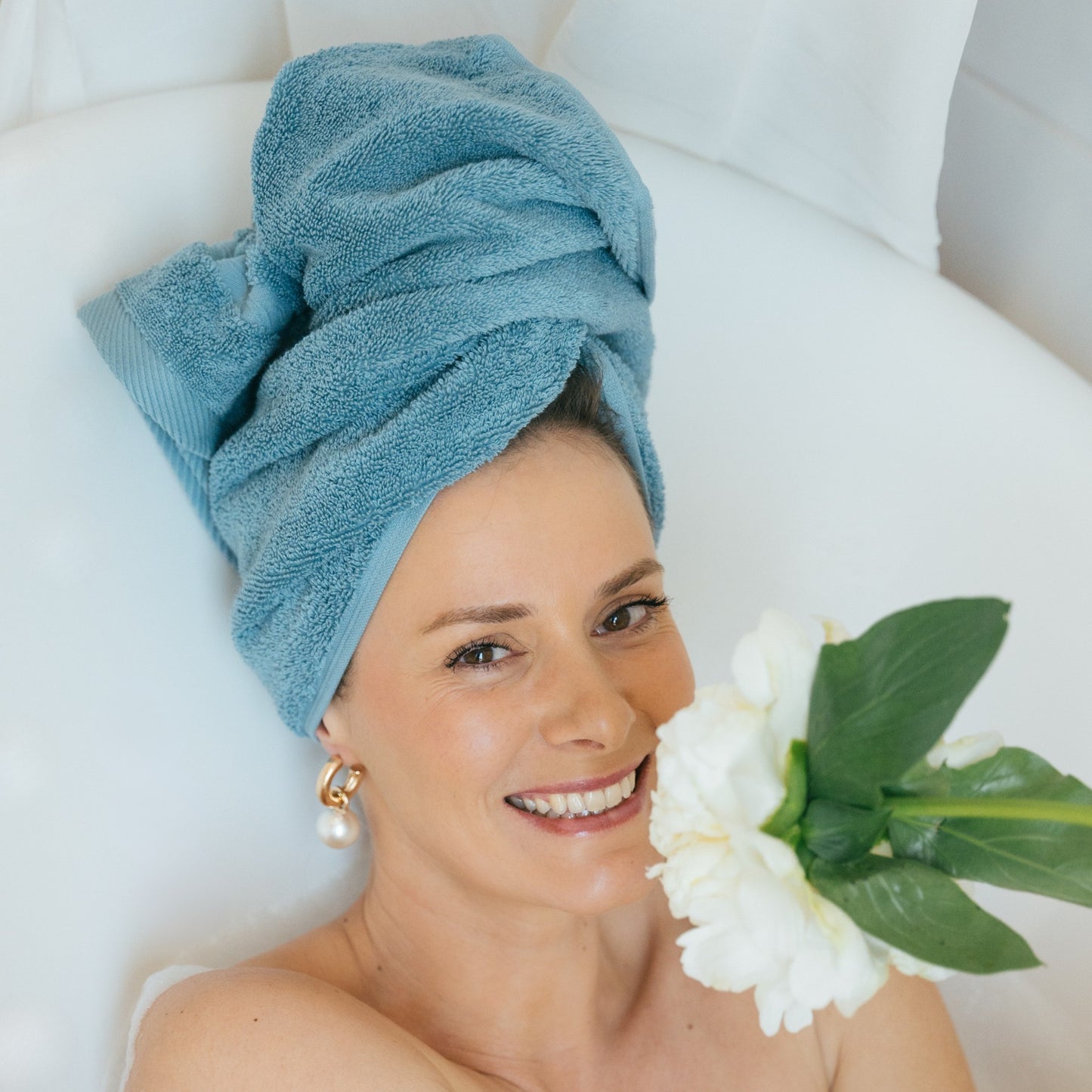 Femme souriante dans un bain moussant, les cheveux enveloppés dans une serviette de toilette FLAMARDA bleu horizon. Ambiance douce et luxueuse avec du linge de bain haut de gamme inspiré de l’océan.