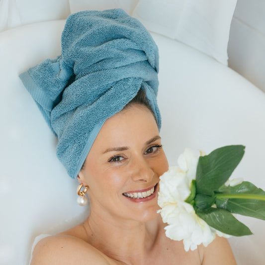 Femme souriante dans un bain moussant, les cheveux enveloppés dans une serviette de toilette FLAMARDA bleu horizon. Ambiance douce et luxueuse avec du linge de bain haut de gamme inspiré de l’océan.