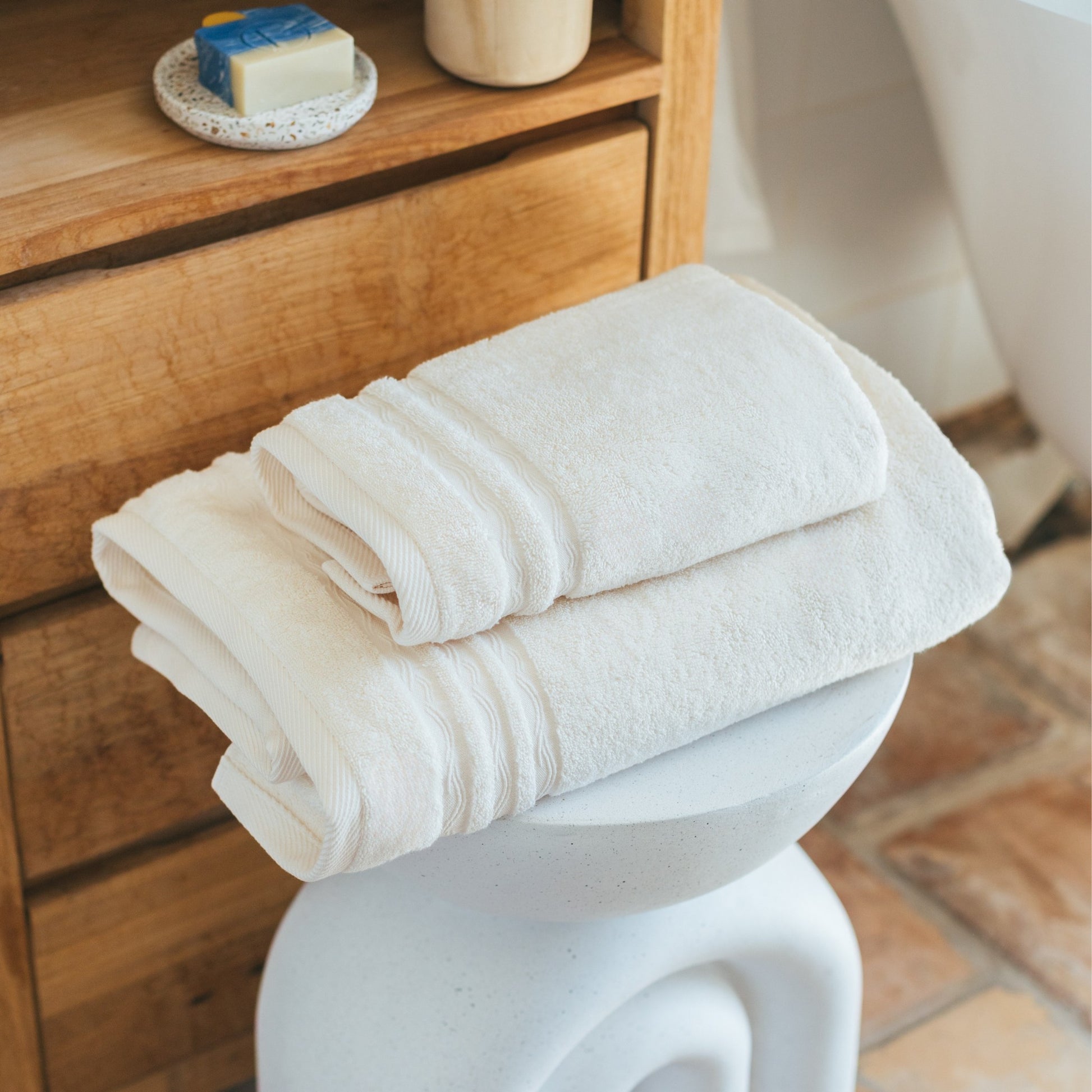 Serviettes de bain FLAMARDA couleur sable naturel posées sur un tabouret design blanc, dans une salle de bain à l’atmosphère douce et chaleureuse. Linge de bain haut de gamme aux tons inspirés des plages portugaises.