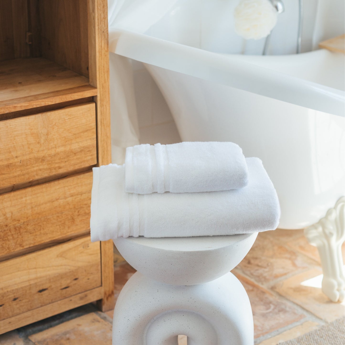 Serviettes de bain FLAMARDA blanches soigneusement pliées sur un tabouret design blanc, dans une salle de bain lumineuse au style épuré. Linge de bain haut de gamme, symbole de pureté, de douceur et d’élégance.