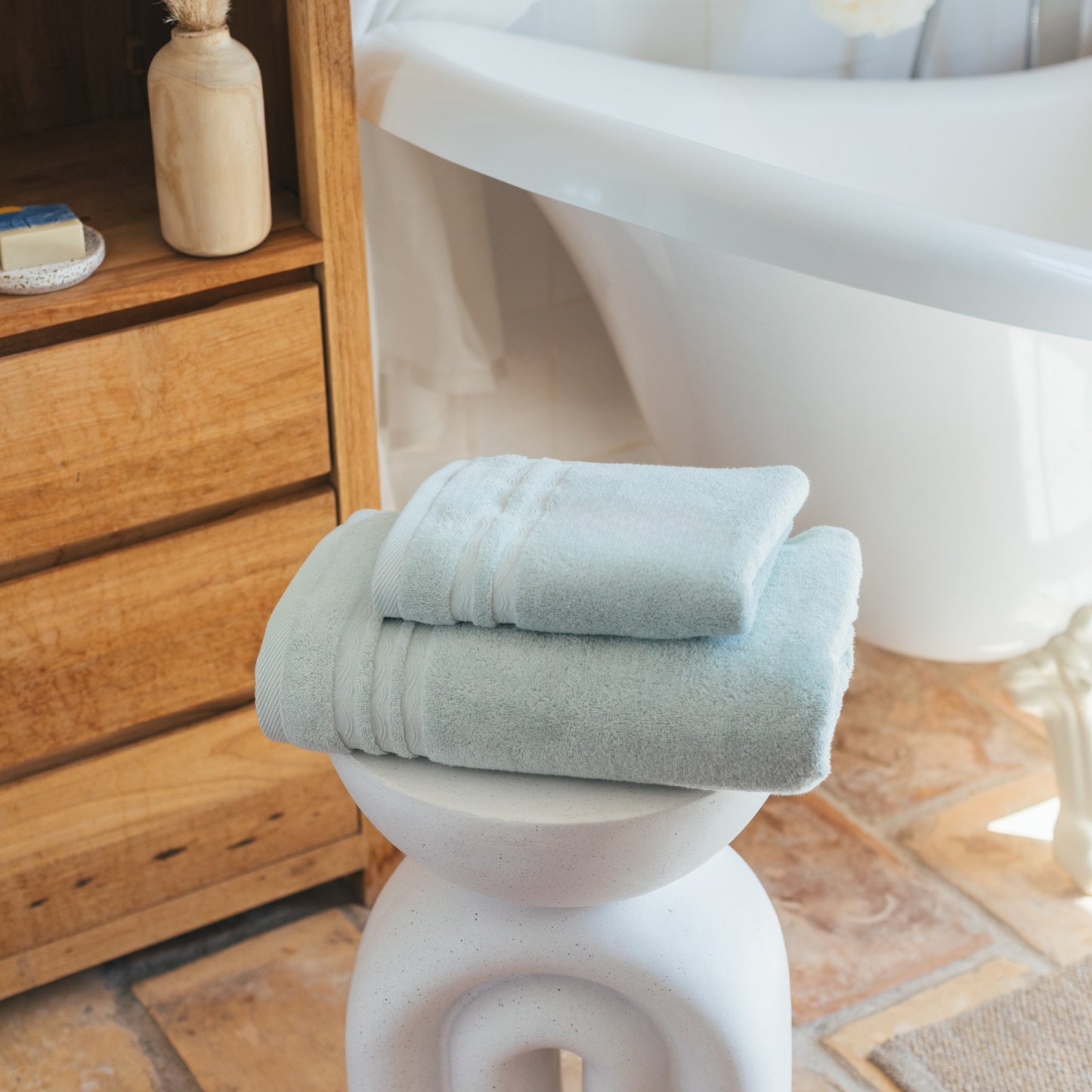 Deux serviettes de bain FLAMARDA vert céladon pliées avec soin sur un tabouret design blanc, dans une salle de bain au style naturel. Linge de bain haut de gamme inspiré de l’océan, aux teintes douces et apaisantes.